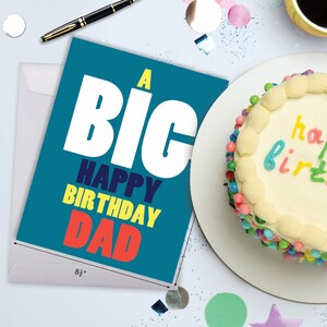 Puede incluir: Una tarjeta azul con las palabras "A Big Happy Birthday Dad" en letras blancas, amarillas y rojas. La tarjeta est&aacute; sobre un sobre blanco. Un pastel con chispas de colores est&aacute; en el fondo.