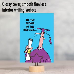 May include: A humorous graduation card with a cartoon illustration of a person wearing a purple graduation gown and cap, holding a diploma and smelling it. The text on the card reads "Ah, the aroma of the diploma..."