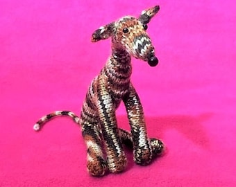 Puede incluir: Un pequeño juguete de perro galgo de punto con rayas marrones y blancas. El juguete está sentado sobre una superficie rosa.