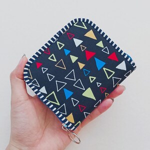 Könnte beinhalten: Ein kleines, quadratisches Portemonnaie mit marineblauem Hintergrund und einem farbenfrohen geometrischen Dreieckmuster. Das Portemonnaie hat einen weiß-marineblauen gestreiften Rand und einen silberfarbenen Reißverschluss.