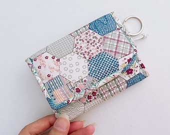 Puede incluir: Una pequeña cartera de tela blanca con un diseño de patchwork que presenta formas hexagonales en tonos azules, rosas y grises. La cartera tiene un llavero.