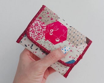 Puede incluir: Una pequeña cartera de tela de patchwork rojo y blanco con ribete rojo. La cartera está doblada por la mitad y se sostiene en una mano.