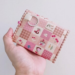 Puede incluir: Una cartera de tela de patchwork rosa y blanca con un peque&ntilde;o anillo de metal. La cartera tiene una variedad de peque&ntilde;os patrones coloridos, incluyendo flores, p&aacute;jaros y casas.