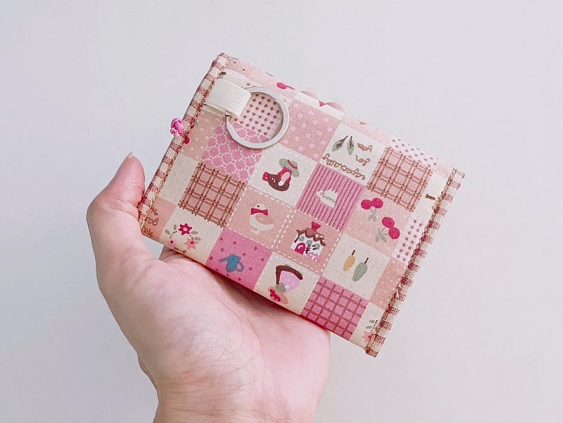Puede incluir: Una cartera de tela de patchwork rosa y blanca con un peque&ntilde;o anillo de metal. La cartera tiene una variedad de peque&ntilde;os patrones coloridos, incluyendo flores, p&aacute;jaros y casas.