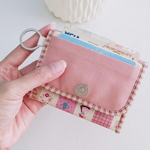 Puede incluir: Una cartera de tela rosa con un estampado floral y un llavero. La cartera tiene un cierre de presi&oacute;n y un peque&ntilde;o bolsillo en la parte delantera.