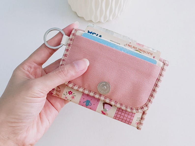 Puede incluir: Una cartera de tela rosa con un estampado floral y un llavero. La cartera tiene un cierre de presi&oacute;n y un peque&ntilde;o bolsillo en la parte delantera.
