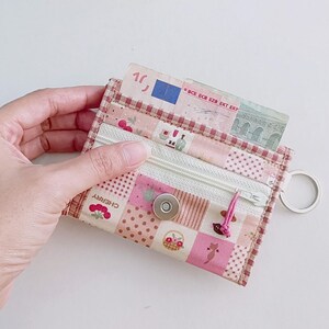 Puede incluir: Una cartera con cremallera de tela de patchwork rosa y blanca con un peque&ntilde;o cierre de presi&oacute;n met&aacute;lico y un llavero. La cartera tiene un estampado floral y de cerezas y el texto "CHERRY" es visible. Un billete de euro es parcialmente visible dentro de la cartera.