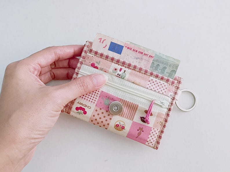 Puede incluir: Una cartera con cremallera de tela de patchwork rosa y blanca con un peque&ntilde;o cierre de presi&oacute;n met&aacute;lico y un llavero. La cartera tiene un estampado floral y de cerezas y el texto "CHERRY" es visible. Un billete de euro es parcialmente visible dentro de la cartera.