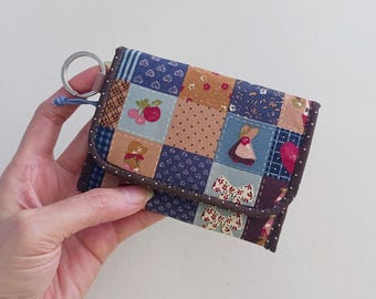 Puede incluir: Una cartera de patchwork con un llavero. La cartera presenta una variedad de cuadrados de tela en tonos azules, marrones y rosas, cada uno con un patrón o diseño diferente, incluyendo cerezas, osos y lazos. La cartera se sostiene en una mano.