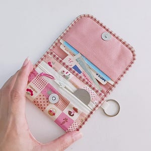 Puede incluir: Una cartera de tela rosa con cierre de cremallera y un llavero. La cartera tiene un dise&ntilde;o de patchwork con una variedad de patrones, incluyendo lunares, cuadros vichy e estampados florales. La cartera est&aacute; abierta y muestra una tarjeta de cr&eacute;dito y una moneda en su interior.