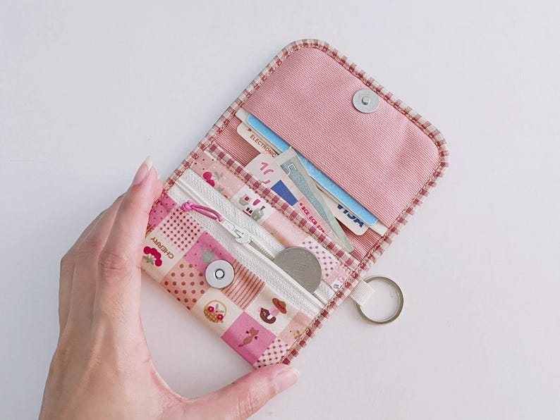 Puede incluir: Una cartera de tela rosa con cierre de cremallera y un llavero. La cartera tiene un dise&ntilde;o de patchwork con una variedad de patrones, incluyendo lunares, cuadros vichy e estampados florales. La cartera est&aacute; abierta y muestra una tarjeta de cr&eacute;dito y una moneda en su interior.