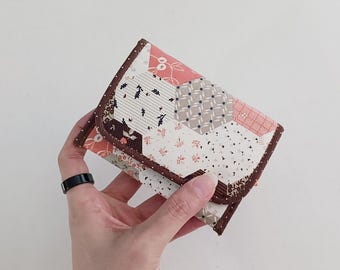 May include: A small, brown and white fabric wallet with a patchwork design featuring hexagonal shapes. The wallet is folded closed and has a brown trim.
