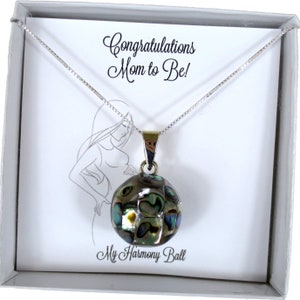 May include: A silver necklace with a round, iridescent pendant, presented in a white gift box. The pendant has a mosaic-like design with shades of green, blue, and brown. The card inside reads "Congratulations Mom to Be!" and "My Harmony Ball".