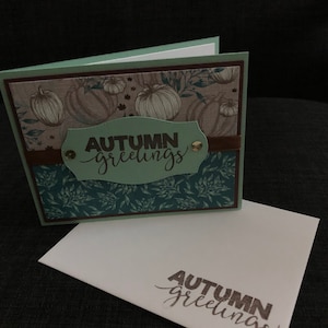 May include: A green card with a brown border and a white envelope. The card has a pattern of pumpkins and leaves. The text "Autumn Greetings" is written in brown on the card. The envelope has the same text written in brown.