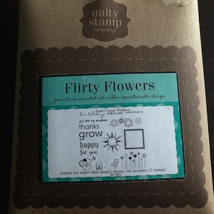 May include: A brown paper package with a teal blue label that says "Flirty Flowers" and "pre-cut un-mounted red rubber repositionable stamps". The label also includes a sample of the stamps with the text "birthday", "HELLO!", "you are my sunshine", "thanks", "grow", "happy", "for you". The package says "we'll change the way you stamp" on the bottom.