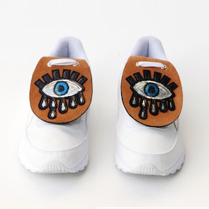 May include: A pair of white sneakers featuring tan leather tongue covers. Each cover is decorated with a beaded eye motif in blue, black, and silver, with black beaded teardrops extending downward from the eye design and white laces.