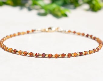 May include: A delicate gold bracelet with small, faceted, reddish-brown gemstones. The bracelet is made of tiny beads and has a simple, elegant design.