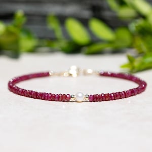 May include: A delicate bracelet made with small red ruby beads and a single white pearl accent. The bracelet is made with gold-toned metal.
