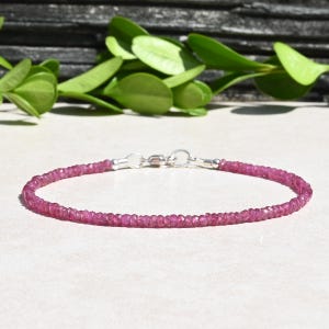 May include: A delicate bracelet featuring small, faceted, pink gemstones strung together. The bracelet is secured with a silver-colored clasp. The background includes green foliage and a light-colored surface.