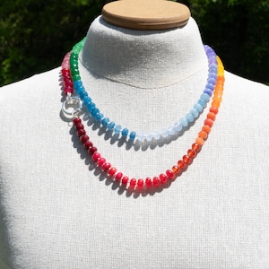 May include: A double-strand necklace displayed on a mannequin. The necklaces feature a gradient of colorful beads, transitioning from red to orange, yellow, green, blue, and purple. A silver-colored clasp connects the strands. The background is blurred.