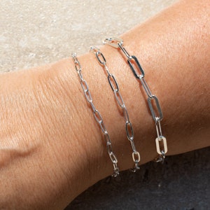 May include: Three silver chain bracelets of different lengths, stacked on a person's wrist.