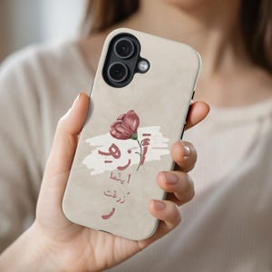 Könnte beinhalten: Beigefarbene Handyhülle mit einem weinroten Blumendesign und arabischer Schrift. Die Hülle hat eine matte Oberfläche und einen Ausschnitt für die Dual-Kamera. Das florale Design wird von einer weißen wolkenartigen Form begleitet.