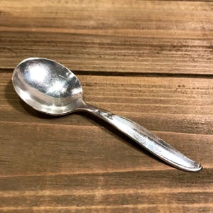 May include: A silver spoon with a rounded bowl and a simple, elegant design. The spoon is lying on a wooden surface.