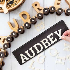 Peut inclure: Un train jouet en bois avec le nom "AUDREY" &eacute;pell&eacute; sur des wagons individuels. Le train est sur une surface blanche avec une pancarte noire qui dit "AUDREY" en lettres blanches. Il y a aussi des lettres en bois blanches &eacute;parpill&eacute;es autour du train et de la pancarte.