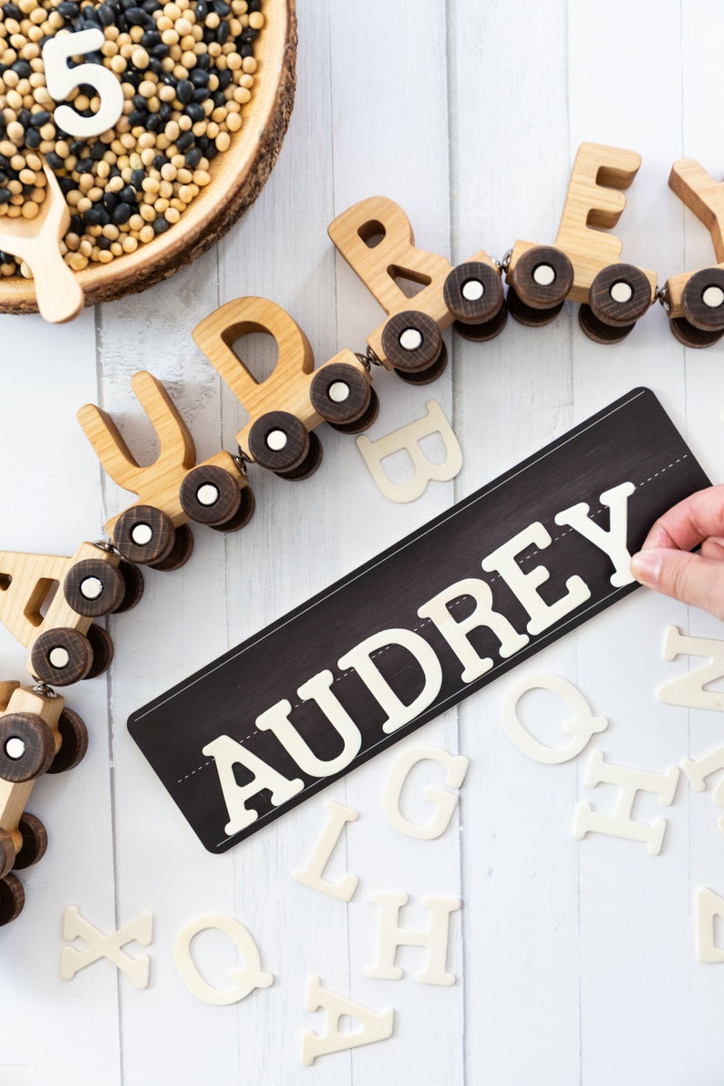 Peut inclure: Un train jouet en bois avec le nom "AUDREY" &eacute;pell&eacute; sur des wagons individuels. Le train est sur une surface blanche avec une pancarte noire qui dit "AUDREY" en lettres blanches. Il y a aussi des lettres en bois blanches &eacute;parpill&eacute;es autour du train et de la pancarte.