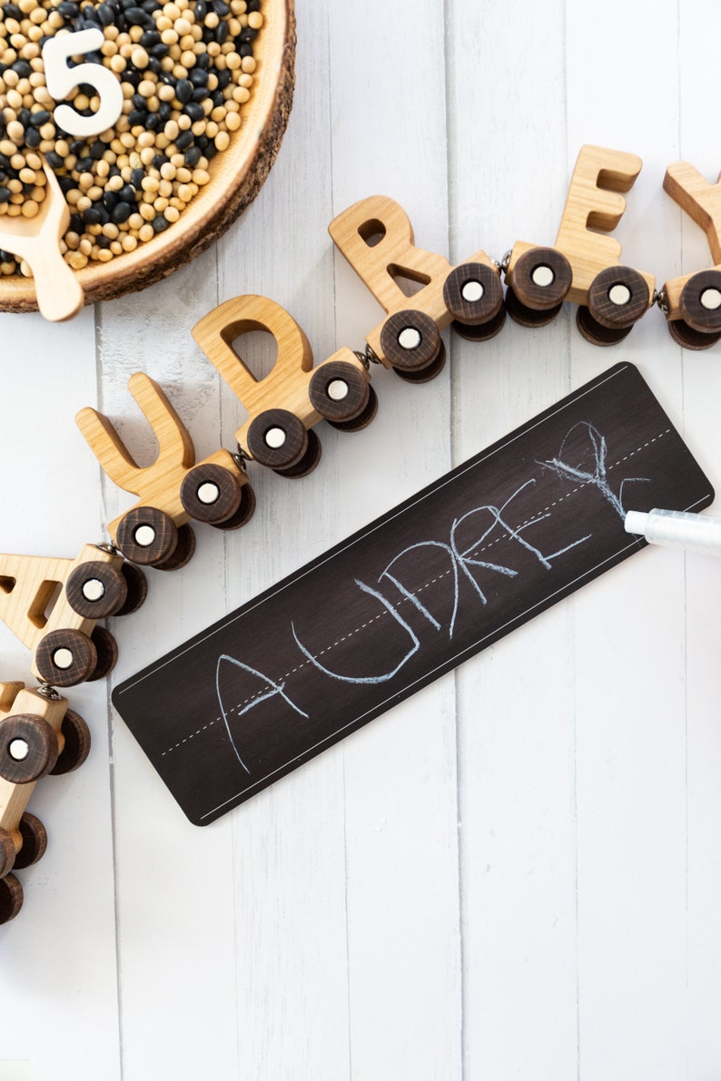 Peut inclure: Train en bois jouet avec les lettres AUDREY sur chaque wagon. Le train est sur une surface blanche avec un tableau noir qui dit AUDREY &eacute;crit dessus &agrave; la craie blanche.