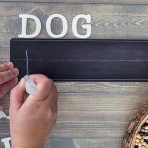 Peut inclure: Une personne &eacute;crit le mot "DOG" sur un tableau noir avec des lettres blanches. Les lettres sont dispos&eacute;es en rang&eacute;e au-dessus du tableau noir. Le tableau noir a une ligne blanche en haut et une ligne blanche en bas. La personne utilise un marqueur blanc pour &eacute;crire le mot.