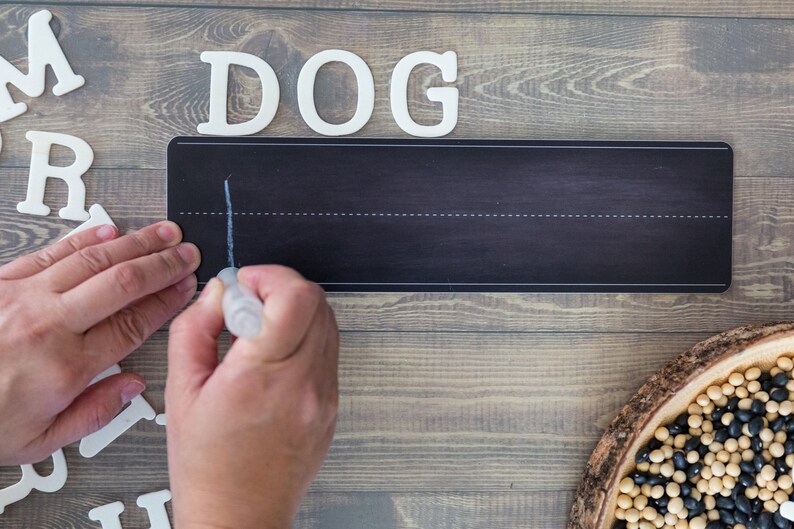 Peut inclure: Une personne &eacute;crit le mot "DOG" sur un tableau noir avec des lettres blanches. Les lettres sont dispos&eacute;es en rang&eacute;e au-dessus du tableau noir. Le tableau noir a une ligne blanche en haut et une ligne blanche en bas. La personne utilise un marqueur blanc pour &eacute;crire le mot.
