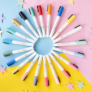 Op de afbeelding: Een cirkelvormige opstelling van kleurrijke stiften met witte barrels en verschillend gekleurde punten en doppen. De achtergrond is roze, blauw en geel, met stervormige decoraties. De stiften zijn voor creatief gebruik.