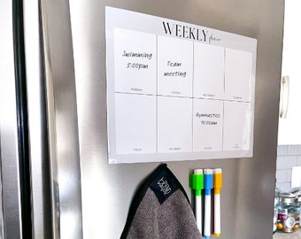 May include: A white dry-erase weekly planner with the word "WEEKLY" at the top. The planner is divided into seven days of the week, with space for writing appointments and tasks. The planner is attached to a stainless steel refrigerator with magnets. There are three dry-erase markers on the refrigerator.