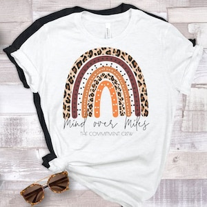 May include: White t-shirt with a brown, orange, and white rainbow design and the text "Mind over Miles" and "The Commitment Crew".