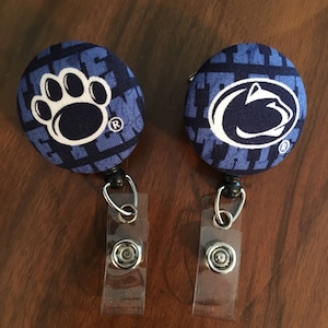 May include: Two blue and white fabric badge holders with a Penn State Nittany Lion logo and a paw print logo. Each holder has a clear plastic retractable badge clip.