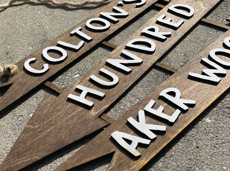 May include: Three wooden arrow signs with white painted lettering. The top sign reads "Colton's", the middle sign reads "Hundred", and the bottom sign reads "Aker Wood".
