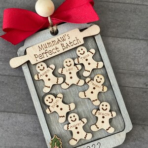 May include: A wooden Christmas ornament shaped like a baking sheet with a rolling pin and gingerbread people. The gingerbread people are decorated with names, including Austin, Levi, Tessa, Rylan, Aubrey, and Gracie. The ornament also includes the text "Mumaw's Perfect Batch" and "2022".