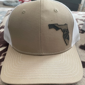May include: A tan and white trucker hat with a black Florida state outline patch. The patch features the Florida state flag and a seal.