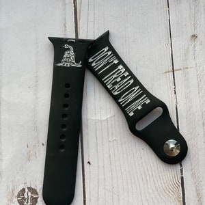May include: Black silicone Apple Watch band with a silver buckle. The band features a white snake with the words "Don't Tread On Me" printed on it.