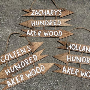 May include: Three wooden arrow signs with white text. The top sign reads "Zachary's Hundred Aker Wood", the middle sign reads "Colten's Hundred Aker Wood", and the bottom sign reads "Holland's Hundred Aker Wood".