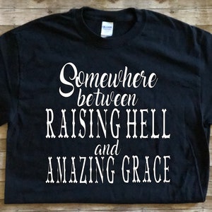 May include: Black t-shirt with white text that reads "Somewhere between raising hell and amazing grace."
