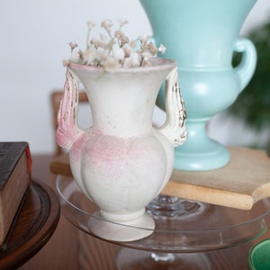May include: A white ceramic vase with pink accents and two handles shaped like hands, filled with white flowers, sits on a clear glass plate on a wooden surface. A light blue ceramic vase with a wide base is behind the white vase.