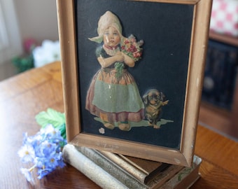 Può includere: Un'illustrazione vintage incorniciata di una ragazza con un vestito blu e bianco che tiene un mazzo di fiori. Un piccolo cane marrone e bianco è in piedi accanto a lei. La cornice è in legno e ha una finitura marrone scuro.