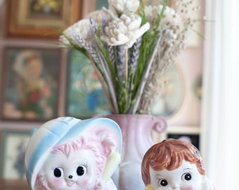 Puede incluir: Dos figuras de cerámica que representan a una criatura rosa y blanca con un sombrero azul y a un niño pequeño de pelo castaño que sostiene un tambor. La criatura está sobre un libro antiguo, mientras que el niño está sentado a su lado.