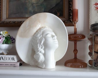 Può includere: Scultura in ceramica bianca di una testa di donna con un grande cappello bianco. La scultura è su uno scaffale con altri oggetti decorativi.