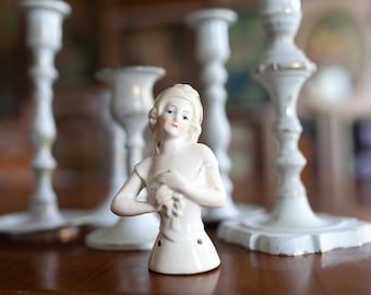 May include: A white porcelain doll bust with painted blue eyes and a delicate floral design on the bodice. The doll is surrounded by white ceramic candle holders.
