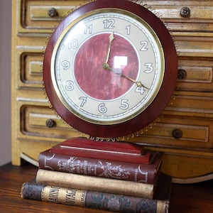 May include: A vintage clock with a red face and gold accents sits on a stack of antique books. The clock has a wooden frame and a decorative metal trim.