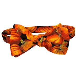 May include: A pre-tied bow tie featuring a vibrant pattern of orange pumpkins with green stems set against a solid black background. The fabric displays multiple pumpkins of varying sizes, creating a dense, repeating autumnal motif across the entire surface.
