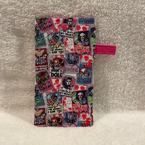 May include: A rectangular fabric case with a colorful collage of horror movie characters and phrases like "You Slay My Heart" and "You Really Float My Boat." The case has a pink button closure and a pink fabric tab.